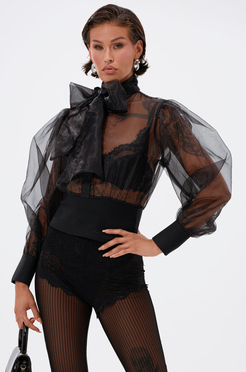 A woman models the DANCE AND SHAKE BLOUSE WITH BOW IN BLACK—sheer with puffed sleeves and a large neck bow—styled with a black corset, lace lingerie, striped tights, and a small handbag against a plain white background.
