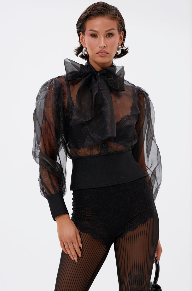 A woman poses in the DANCE AND SHAKE BLOUSE WITH BOW IN BLACK, a sheer organza style with puffed sleeves and a statement neck bow, paired with high-waisted black lace shorts and striped sheer tights against a white background.