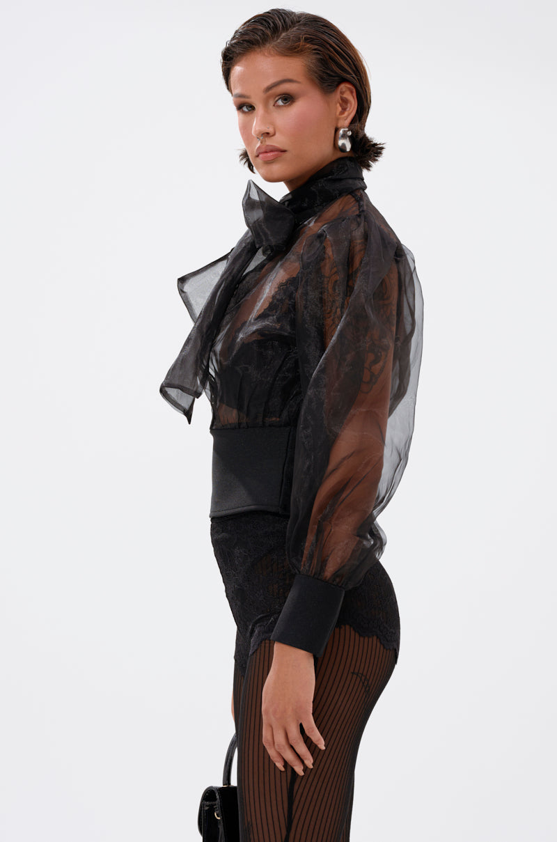 A woman with short, slicked-back hair models the DANCE AND SHAKE BLOUSE WITH BOW IN BLACK, a sheer organza top with puffy sleeves and a large neck bow. She pairs it with striped black tights and poses sideways against a plain white background.