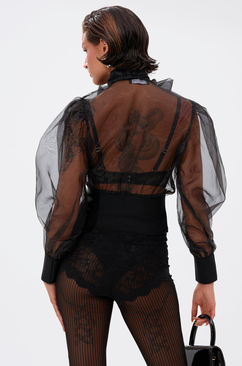 A person with short, slicked-back hair wears the DANCE AND SHAKE BLOUSE WITH BOW IN BLACK— a sheer organza top with puffy sleeves—paired with a visible bra, lacy black bottoms, patterned tights, and carrying a shiny black handbag while facing away.