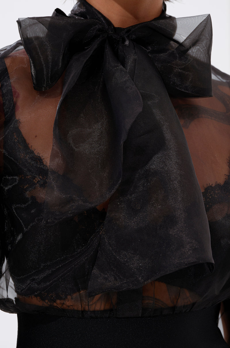 Close-up of the DANCE AND SHAKE BLOUSE WITH BOW IN BLACK—a sheer statement top featuring a large bow at the neck. The black organza reveals a bra underneath, creating a bold and stylish layered effect.