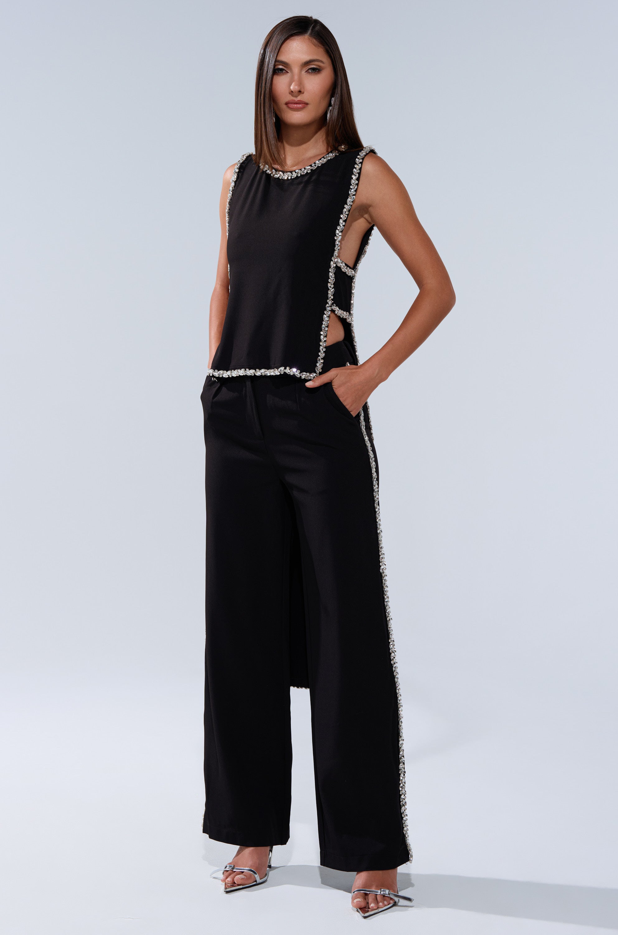 A woman stands confidently in the TALK TO ME BABY BLOUSE WITH RHINESTONE, a sleeveless black top with side cutouts and pearl-like details, paired with wide-leg pants, silver open-toe heels, and straight brown hair.