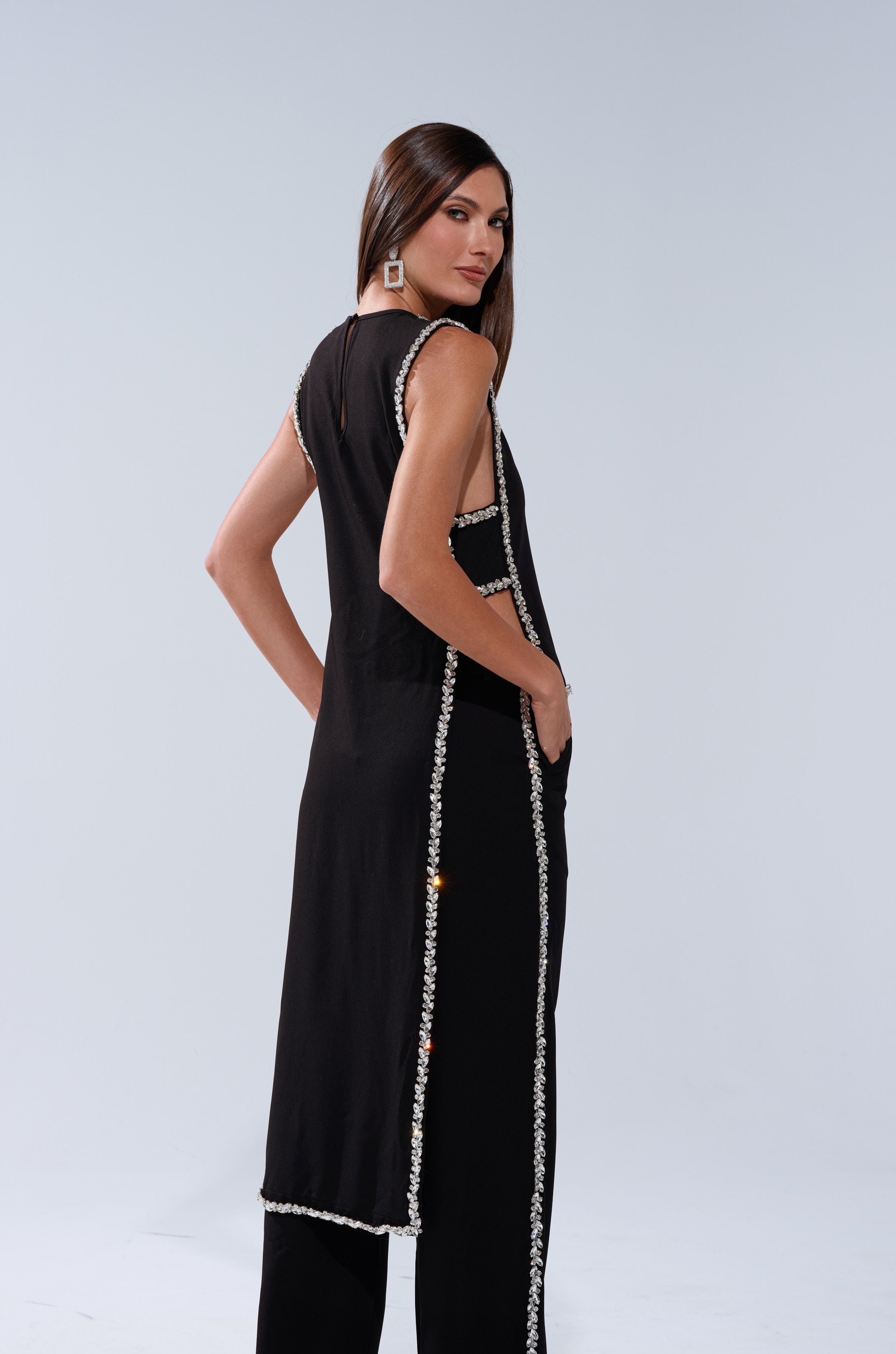 A woman with long brown hair poses sideways against a plain light background, wearing the TALK TO ME BABY BLOUSE WITH RHINESTONE, sleeveless and black with white beaded trim, her hand in her pocket.