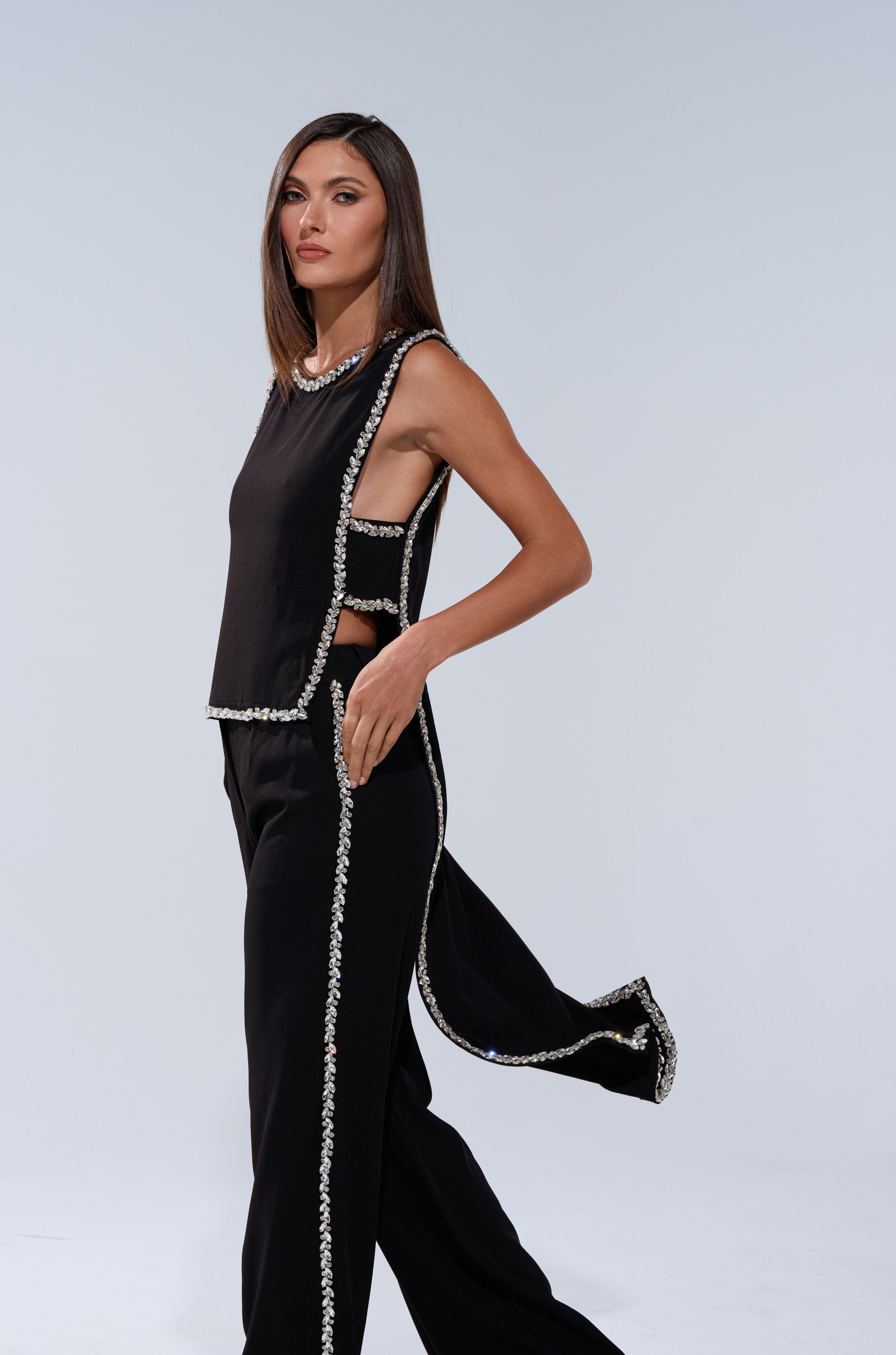 A woman with straight brown hair models the TALK TO ME BABY BLOUSE WITH RHINESTONE, a black sleeveless top adorned with sparkling rhinestone trim, paired with matching pants against a plain light gray background.