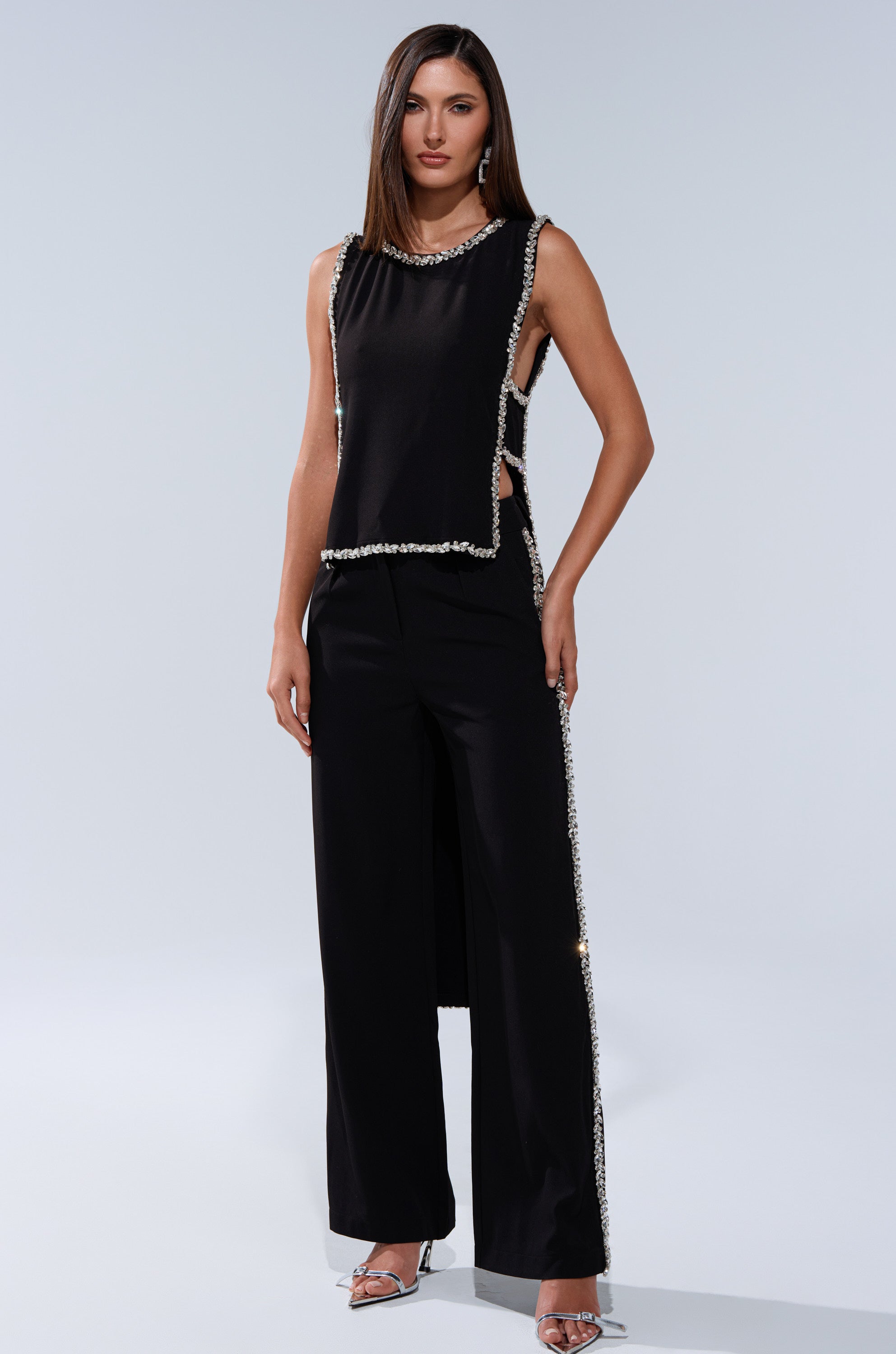A woman stands against a plain light background wearing the TALK TO ME BABY BLOUSE WITH RHINESTONE—a sleeveless black cropped blouse with silver embellishments—paired with matching wide-leg pants and silver high-heeled sandals.