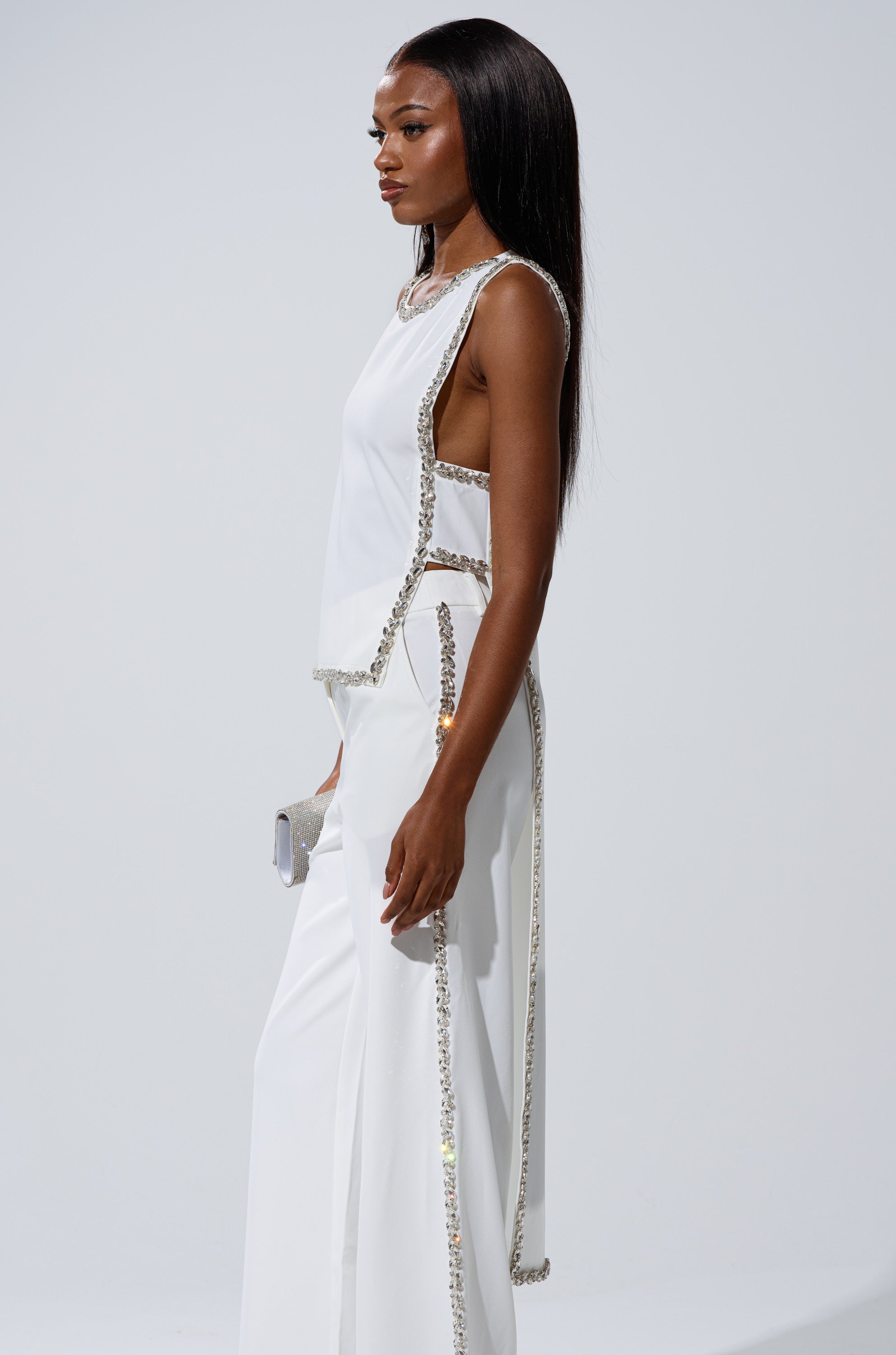 A woman with long straight hair wears the TALK TO ME BABY BLOUSE WITH RHINESTONE IN WHITE and holds a small clutch, standing against a plain light background.