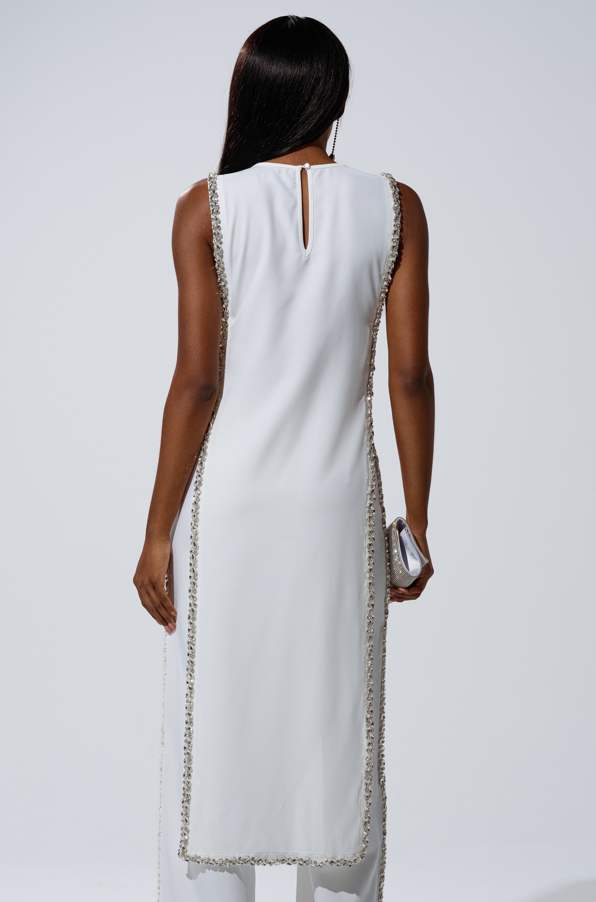 A woman with long dark hair is seen from the back wearing the TALK TO ME BABY BLOUSE WITH RHINESTONE IN WHITE, a sleeveless top with beaded trim. She holds a small silver clutch in her right hand against a plain light background.