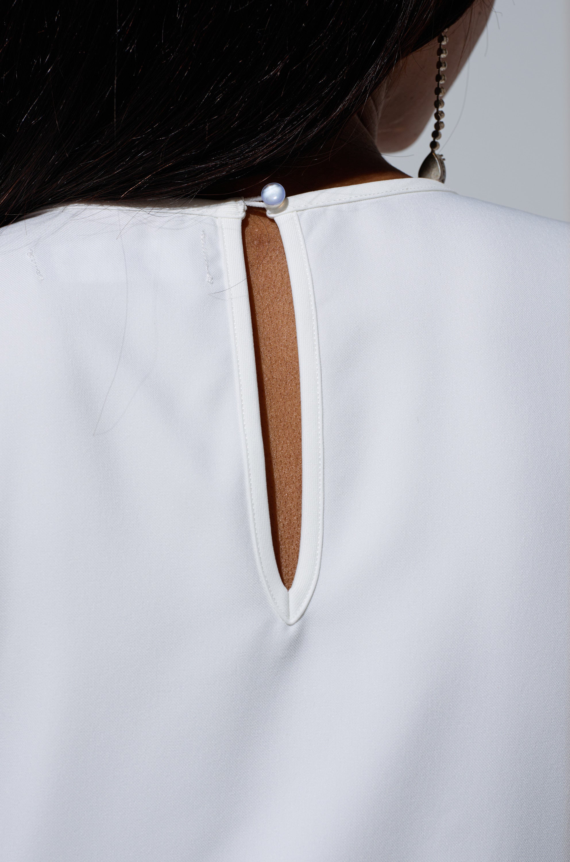 Close-up of the back of the TALK TO ME BABY BLOUSE WITH RHINESTONE IN WHITE, featuring a white sleeveless design with an oval keyhole and pearl button closure. A strand of dark hair and a dangling earring are partially visible.