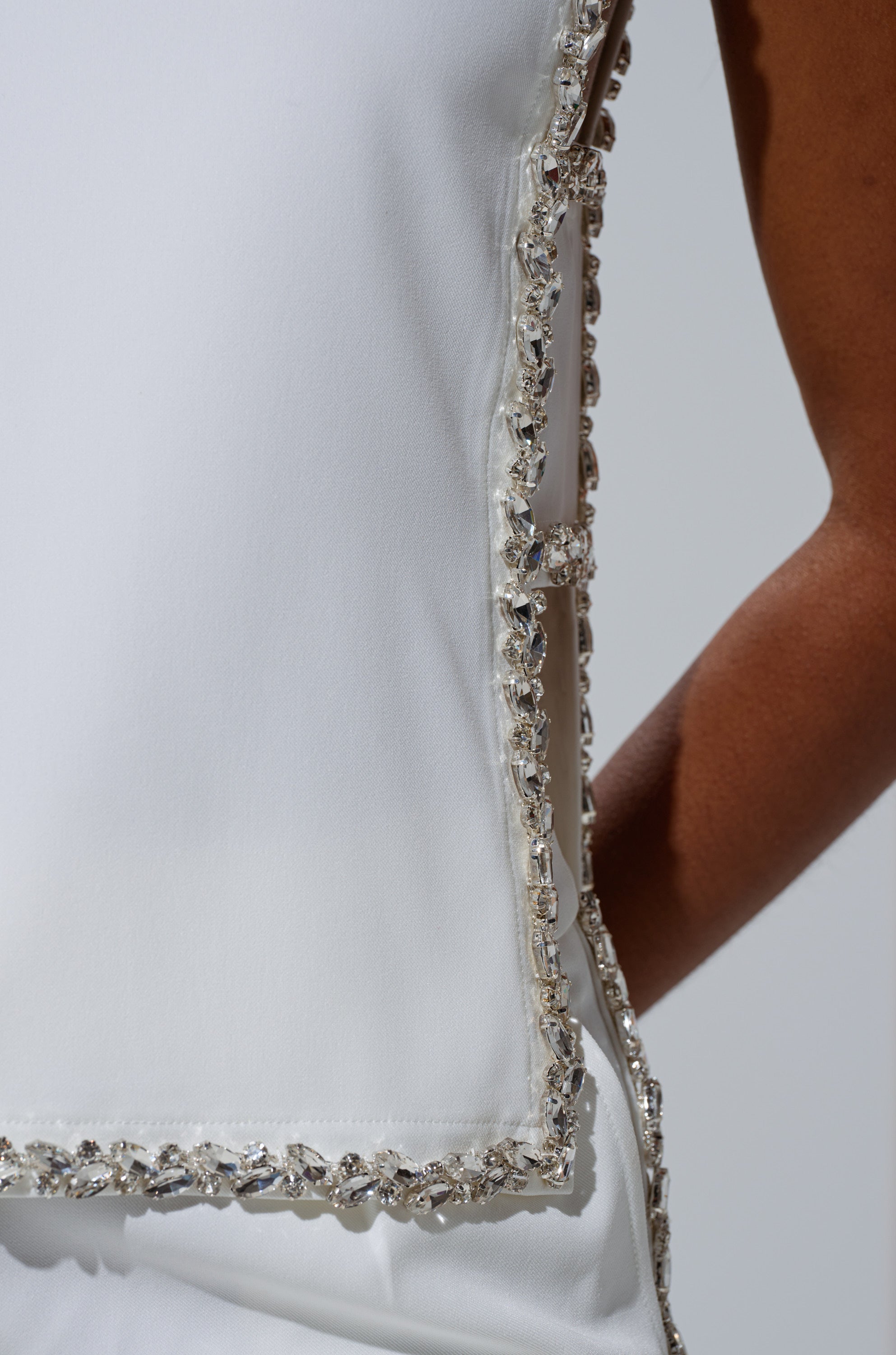 Close-up of the TALK TO ME BABY BLOUSE WITH RHINESTONE IN WHITE, a sleeveless cropped blouse featuring a decorative border of clear rhinestones, worn by a person with medium brown skin; only part of their arm and torso is visible.