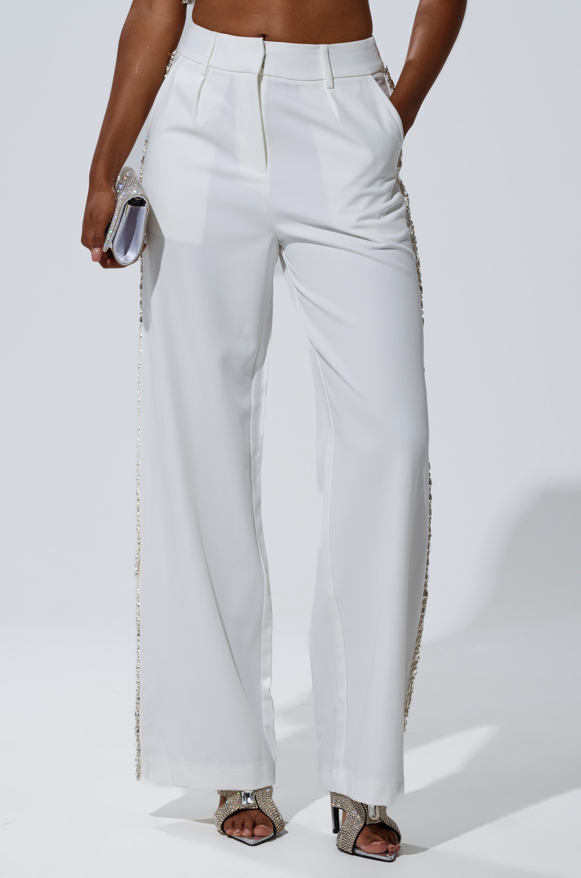 A person wearing the TALK TO ME TROUSER WITH RHINESTONE IN WHITE with hands in pockets, holding a small silver clutch, and open-toe heeled sandals adorned with rhinestones.