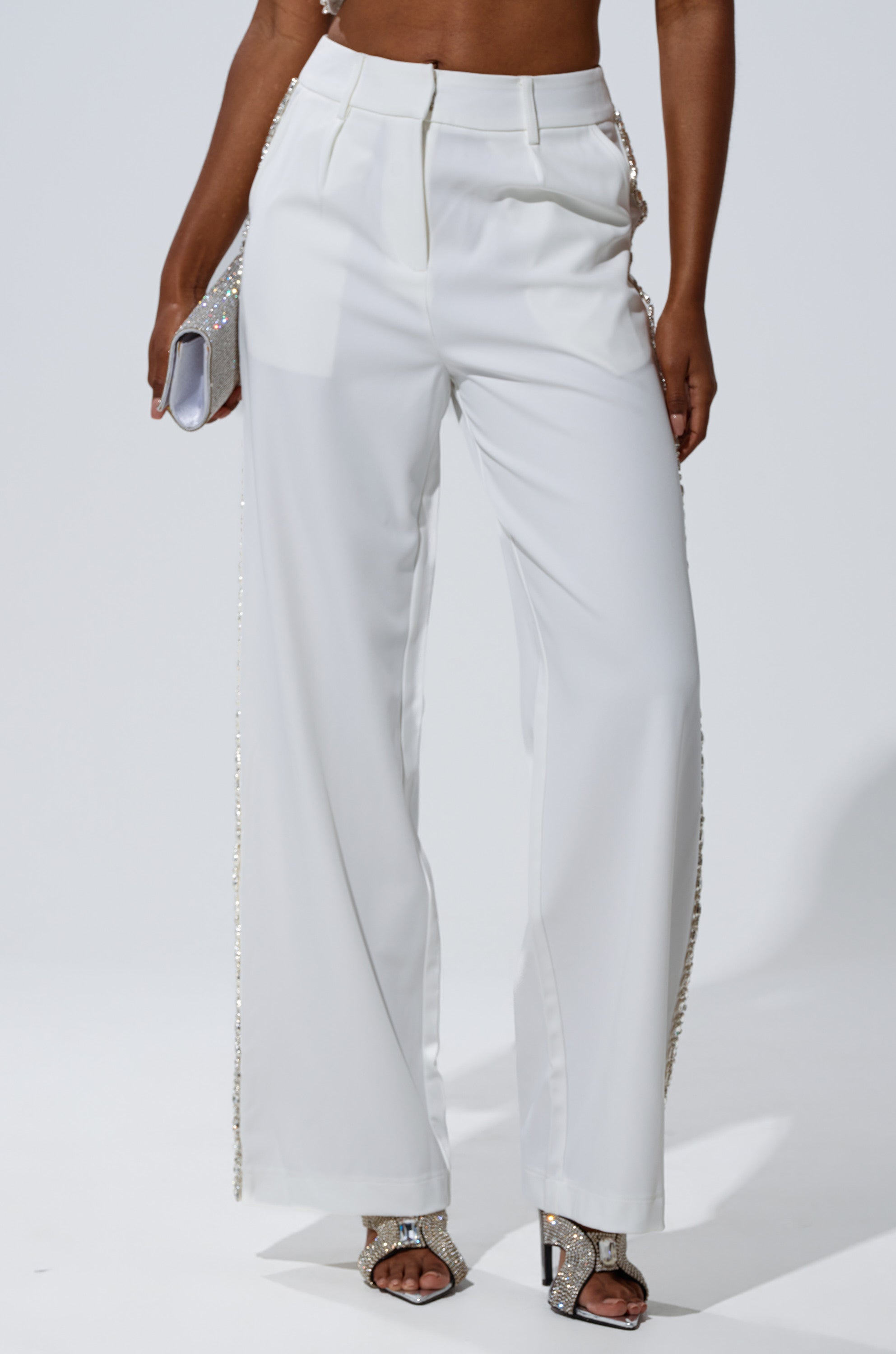 A person wearing TALK TO ME TROUSER WITH RHINESTONE IN WHITE, styled with a silver clutch and silver open-toe heels, stands against a plain light background.
