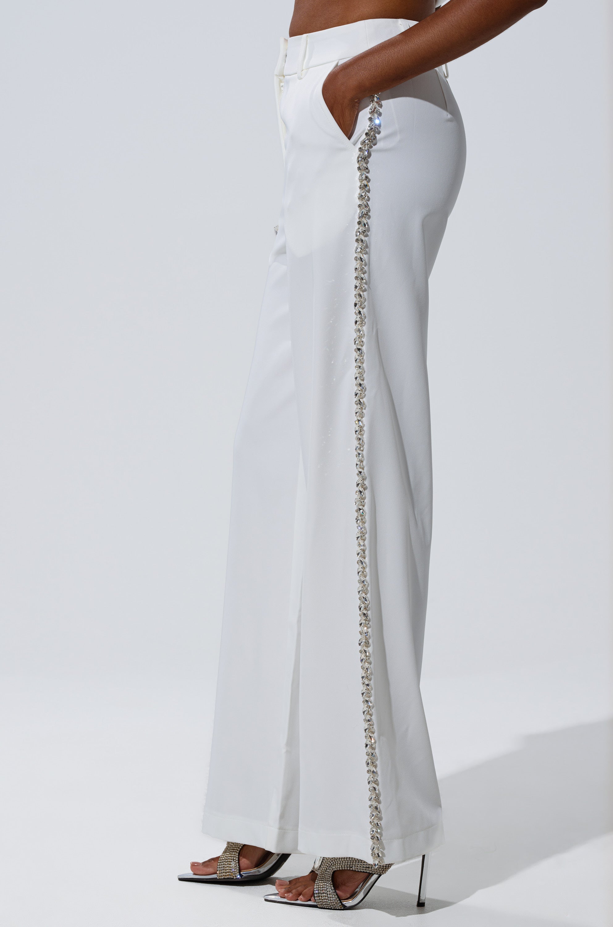 Wearing the TALK TO ME TROUSER WITH RHINESTONE IN WHITE—high-rise, straight-leg pants with rhinestone-trimmed side seams, hands in pockets, styled with open-toe heels.