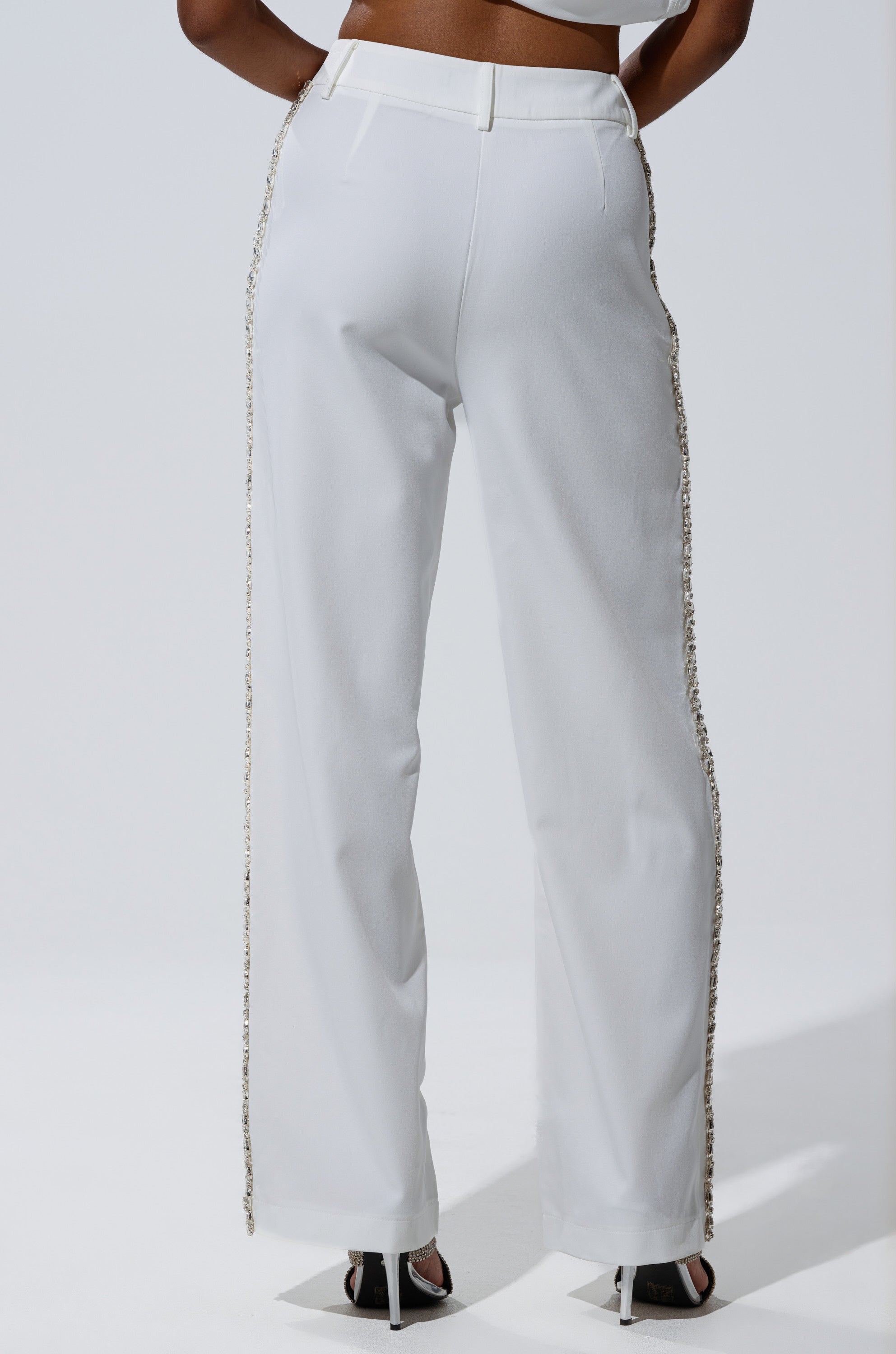 A person stands with their back to the camera, wearing the TALK TO ME TROUSER WITH RHINESTONE IN WHITE—high-rise, straight-leg pants with side rhinestone embellishments and belt loops—paired with black open-toe heels.