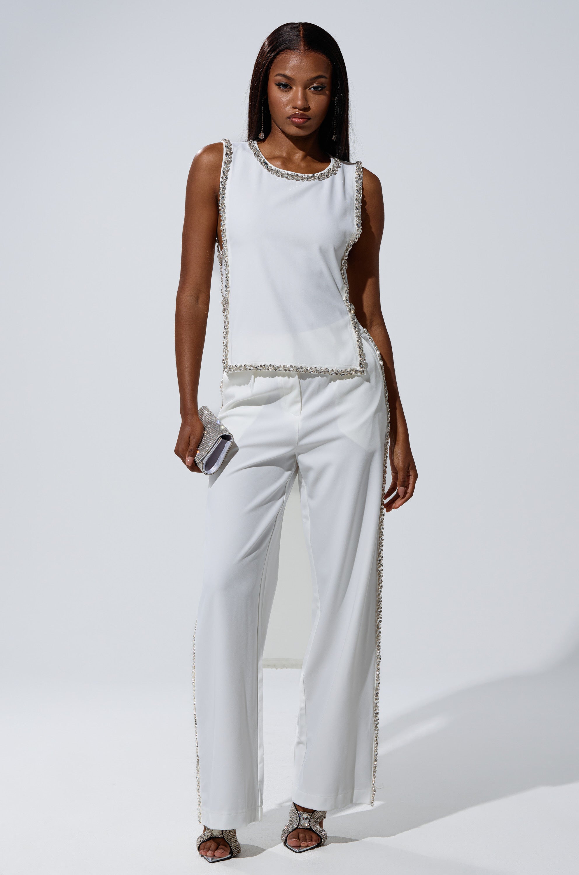 A woman poses confidently against a plain white background, wearing the TALK TO ME TROUSER WITH RHINESTONE IN WHITE, paired with a sleeveless white top, strappy heels, and a small silver clutch.