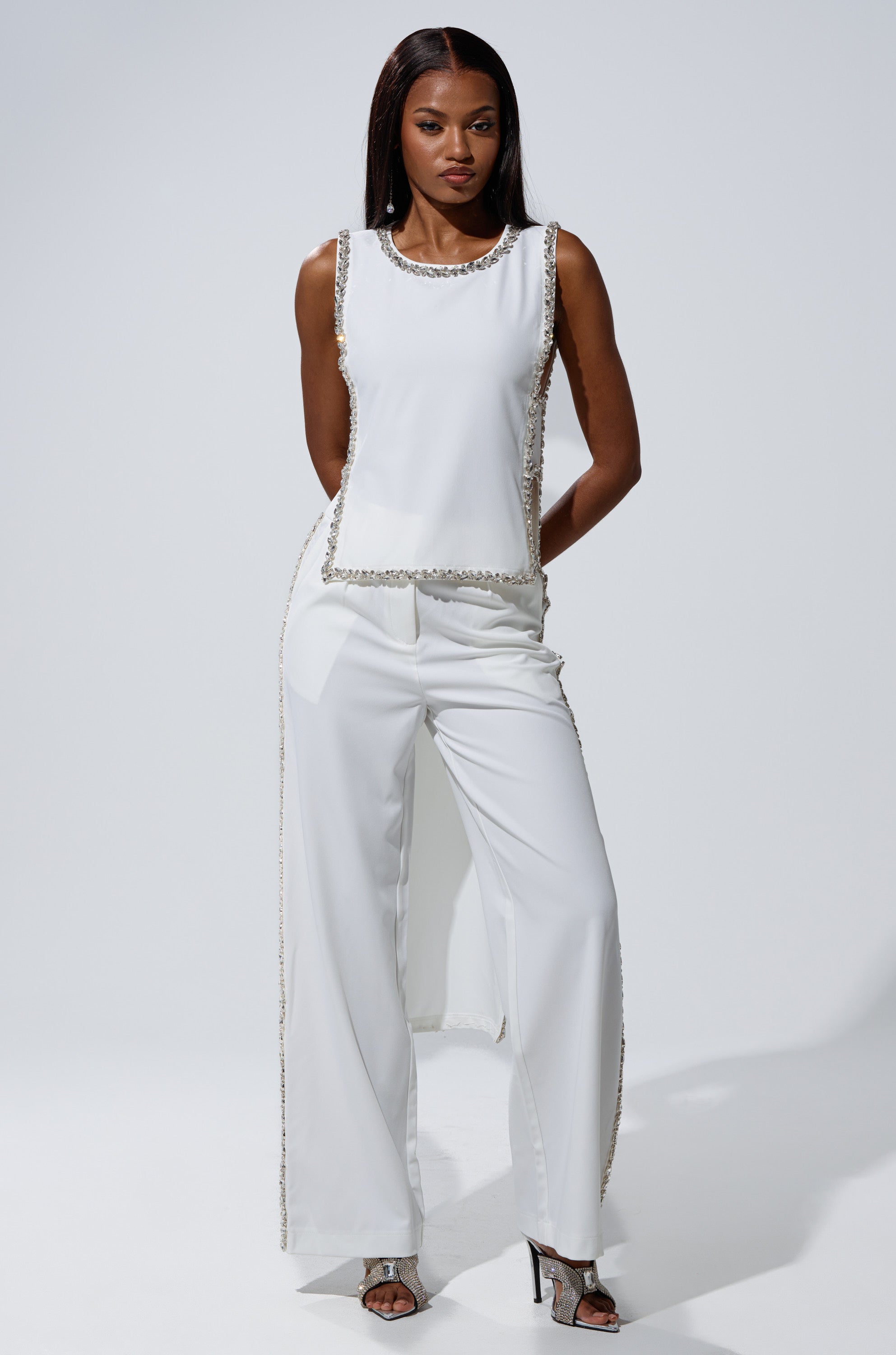 A woman stands confidently against a plain background, wearing the TALK TO ME TROUSER WITH RHINESTONE IN WHITE—a high-rise, straight-leg pant—paired with a sleeveless white tunic and open-toe heels. Her hands are behind her back.