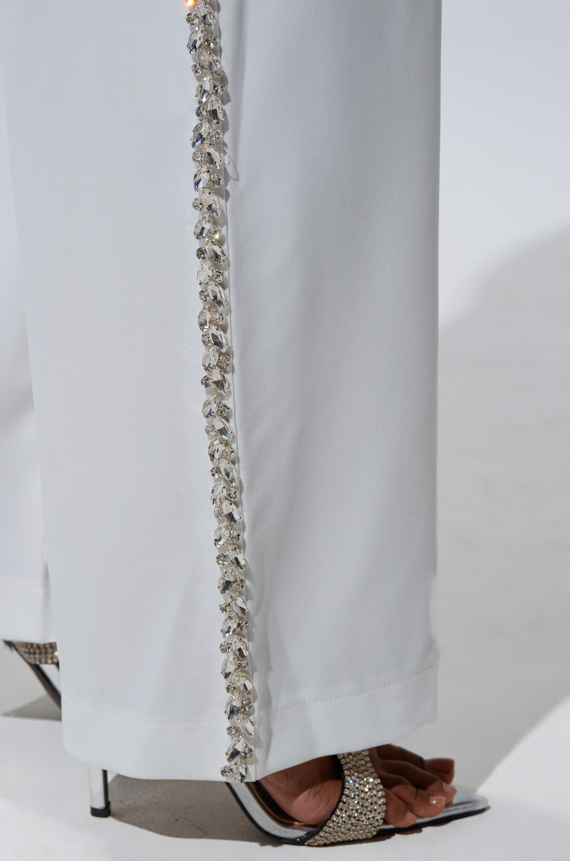 A close-up of the TALK TO ME TROUSER WITH RHINESTONE IN WHITE, a high-rise straight-leg pant with silver and crystal rhinestone trim, styled with sparkling open-toe heels.