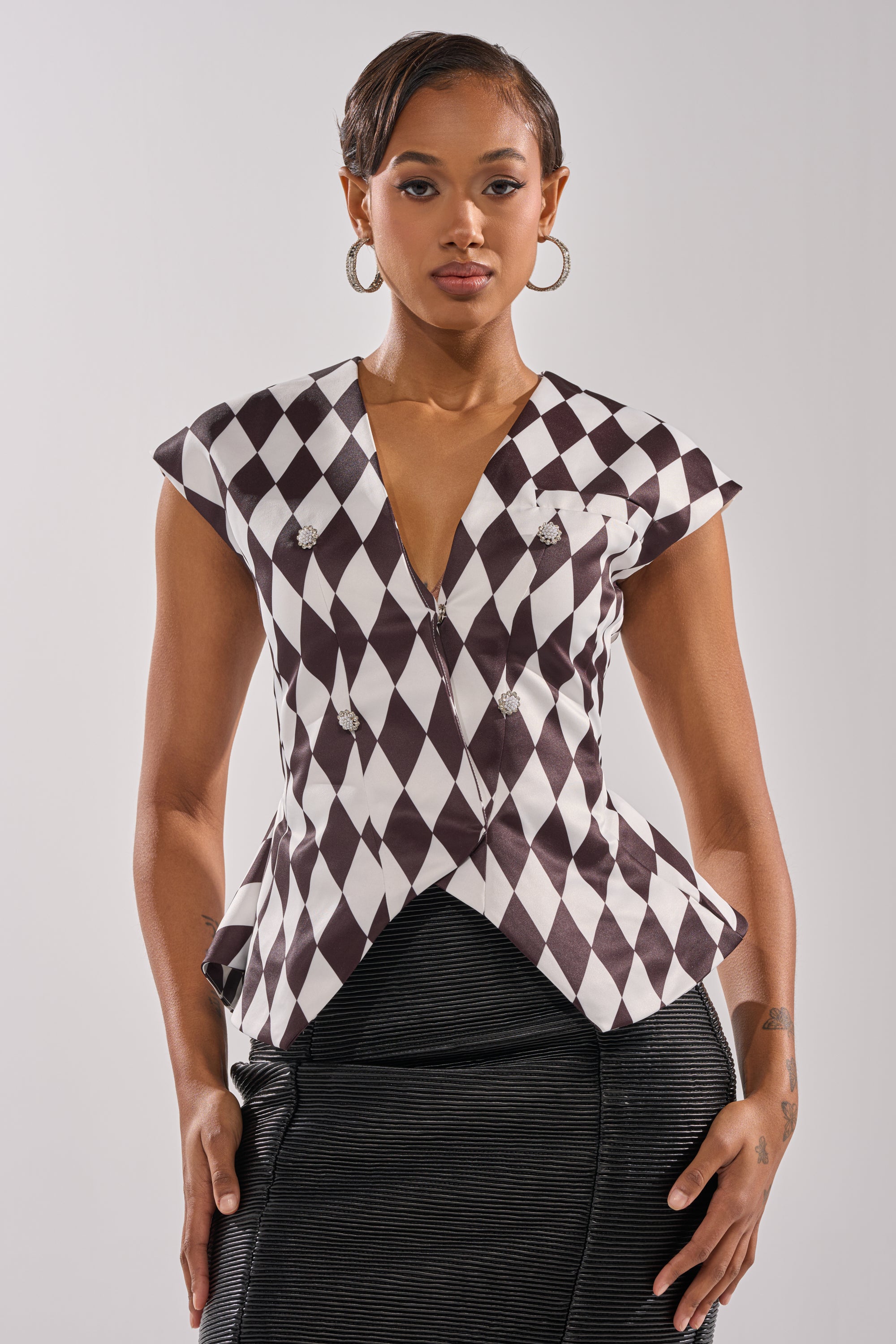 A woman faces forward, wearing the GUESS WHO V-NECK BLOUSE featuring a black-and-white diamond pattern and silver buttons, paired with a dark textured skirt. She accessorizes with hoop earrings and has slicked-back hair.