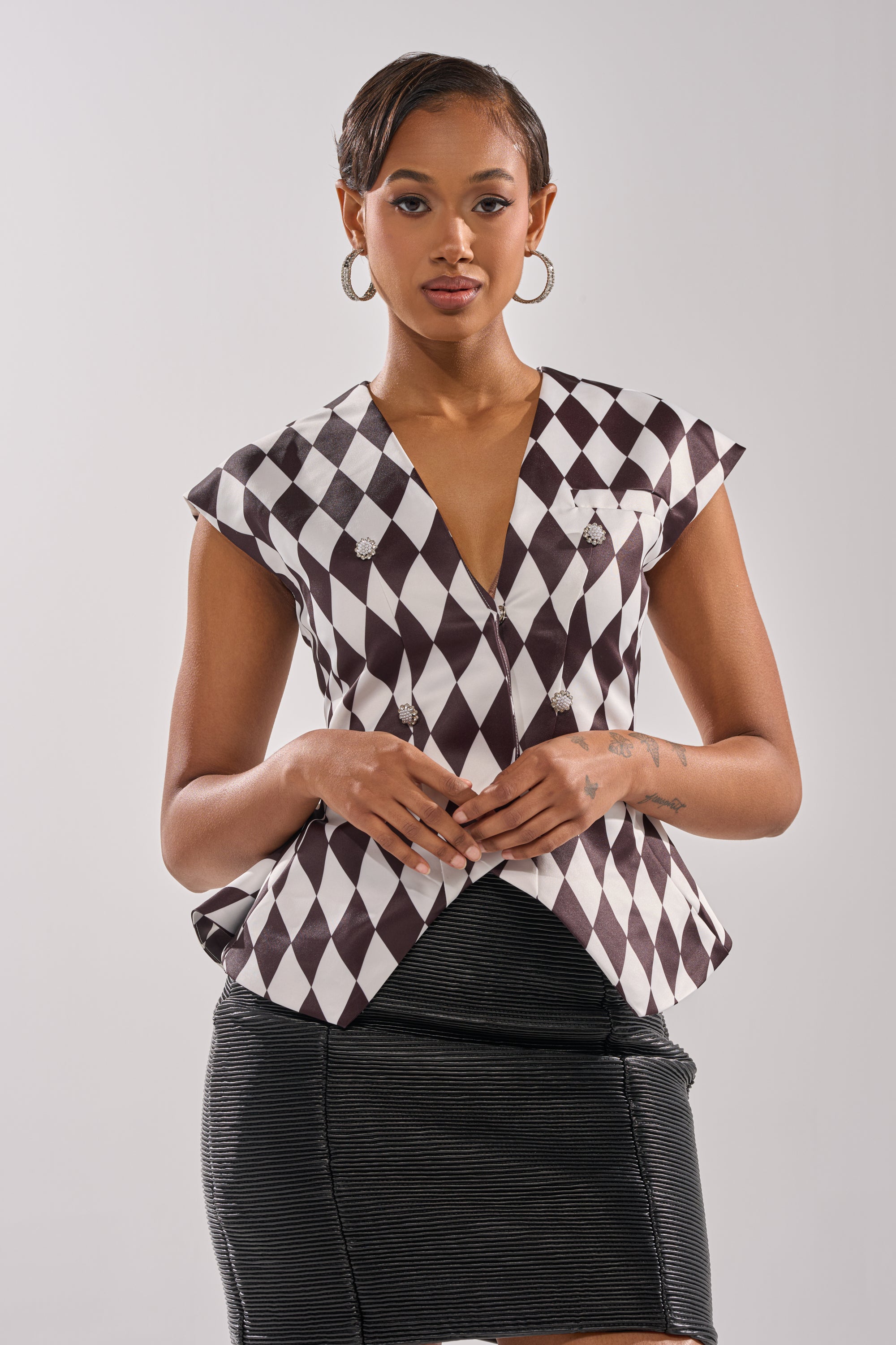A woman stands against a plain background, wearing the GUESS WHO V-NECK BLOUSE with a black and white diamond pattern, short sleeves, and deep neckline, paired with a textured black skirt. She accessorizes with hoop earrings and has neatly styled hair.