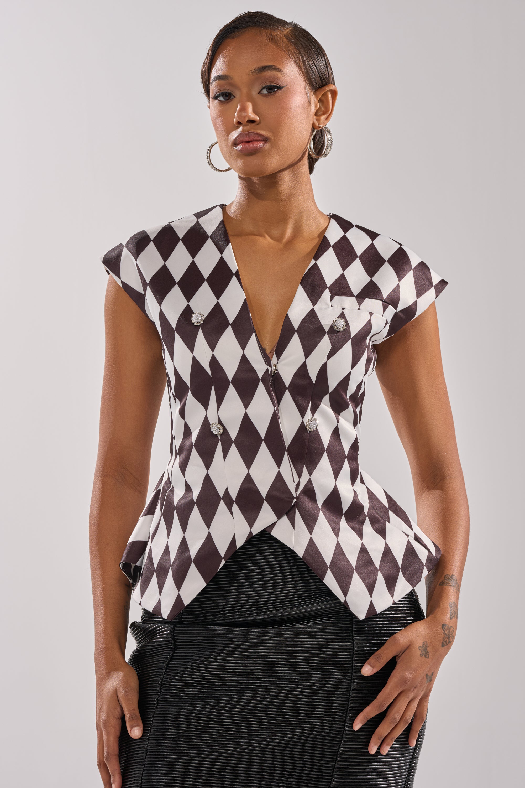 A woman with short hair and hoop earrings wears the GUESS WHO V-NECK BLOUSE, featuring a black-and-white diamond pattern and cap sleeves in satin-like fabric, paired with a dark skirt against a plain light background.