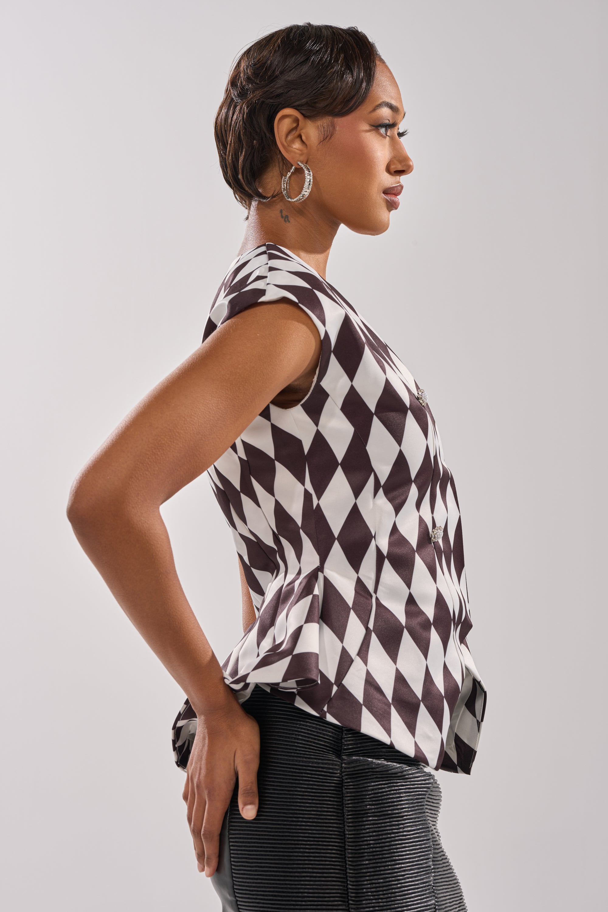 A woman stands in profile with her hands on her hips, wearing the GUESS WHO V-NECK BLOUSE in black and white satin diamond pattern, paired with a dark skirt and hoop earrings, against a plain light background.