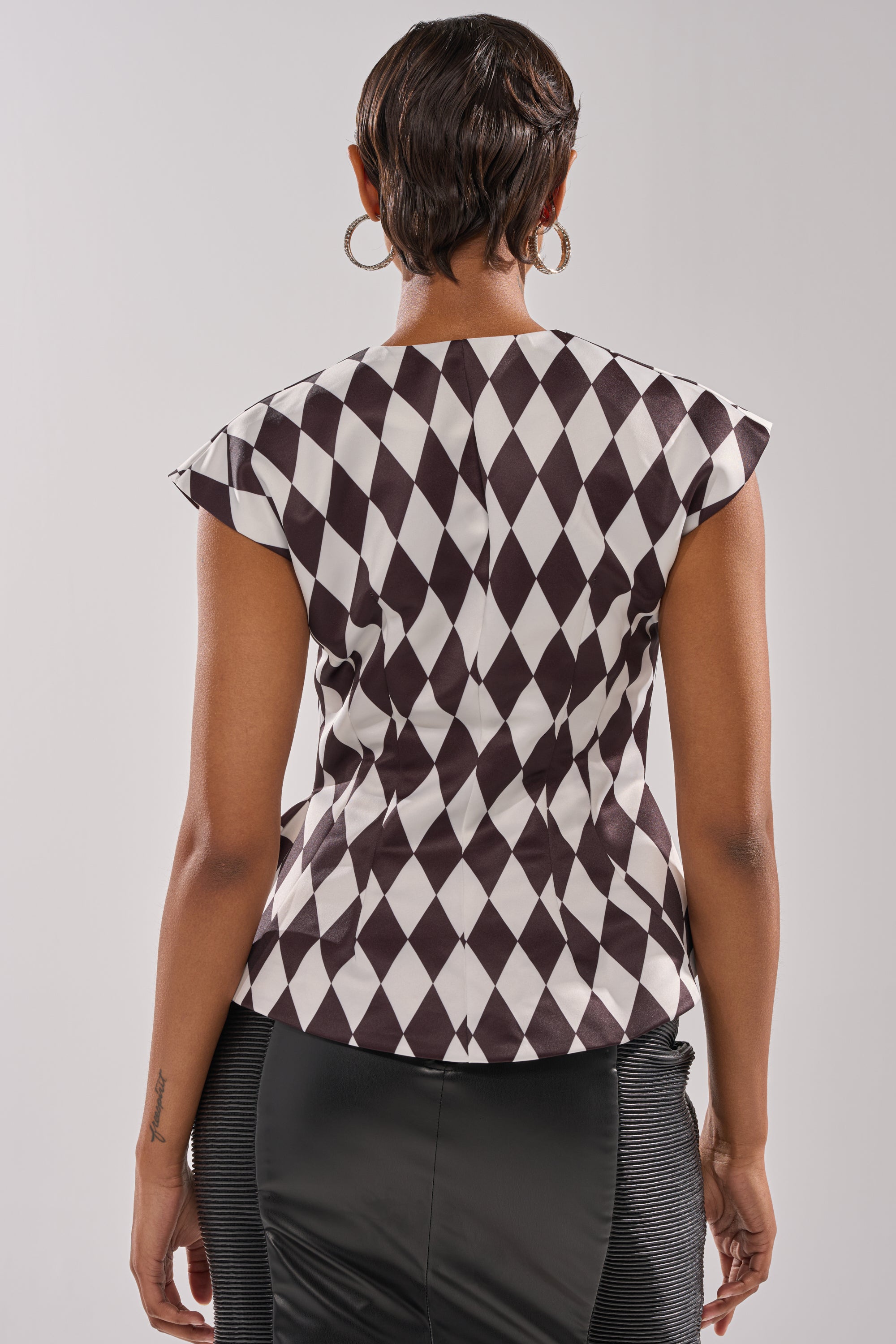 A person with short hair and large hoop earrings is seen from the back against a plain light background, wearing the GUESS WHO V-NECK BLOUSE with a black and white diamond pattern, paired with a black skirt.