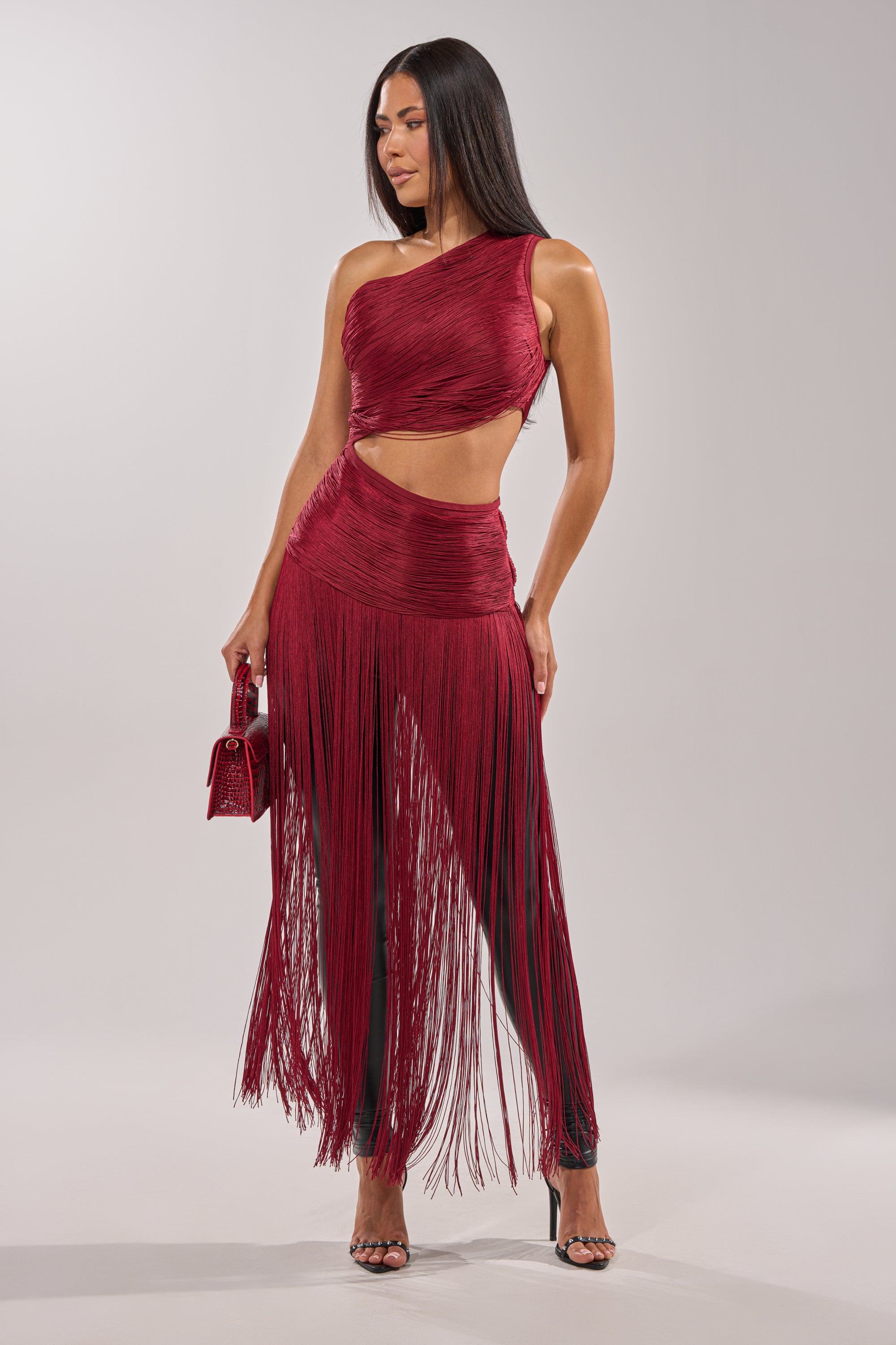 A woman stands confidently in the LOVE STRUCK FRINGE TOP, a red one-shoulder crop top with long fringe detailing. She accessorizes with a small red handbag and strappy high heels, her long hair down against a plain background. 1
