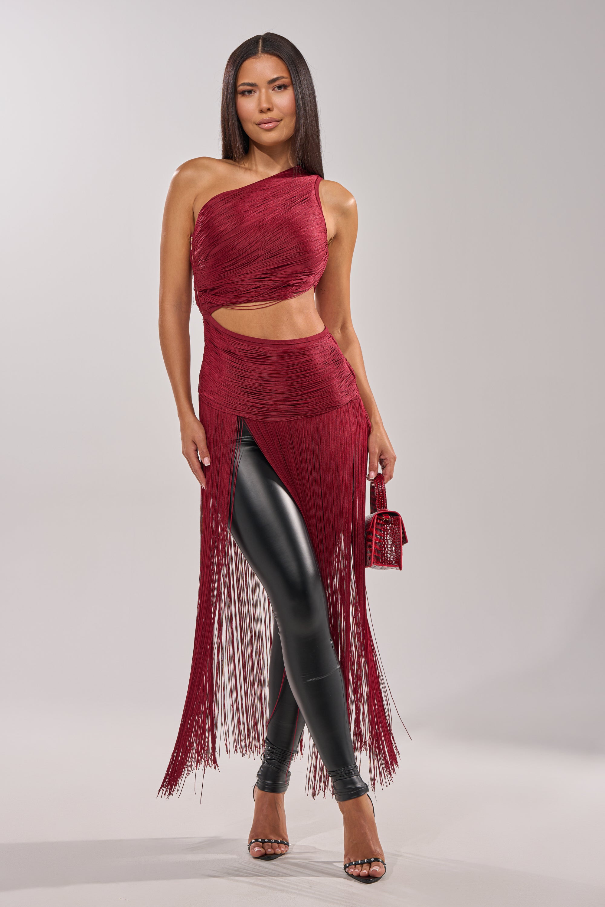 A woman stands against a plain background wearing the LOVE STRUCK FRINGE TOP in burgundy, styled with black leather leggings, strappy heels, and a small matching burgundy handbag. 2