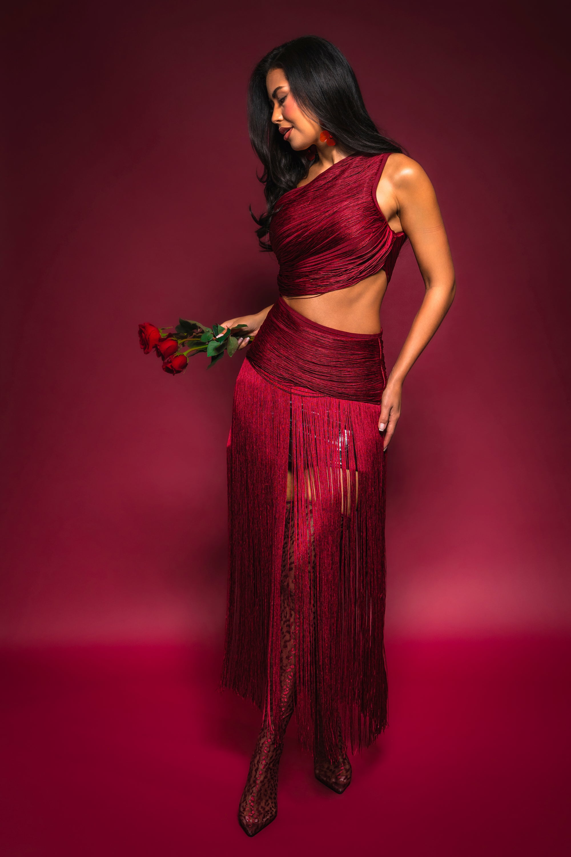 Wearing the LOVE STRUCK FRINGE TOP, a woman holds three red roses against a matching backdrop, her gentle smile and the top’s asymmetrical fringe design capturing effortless elegance.