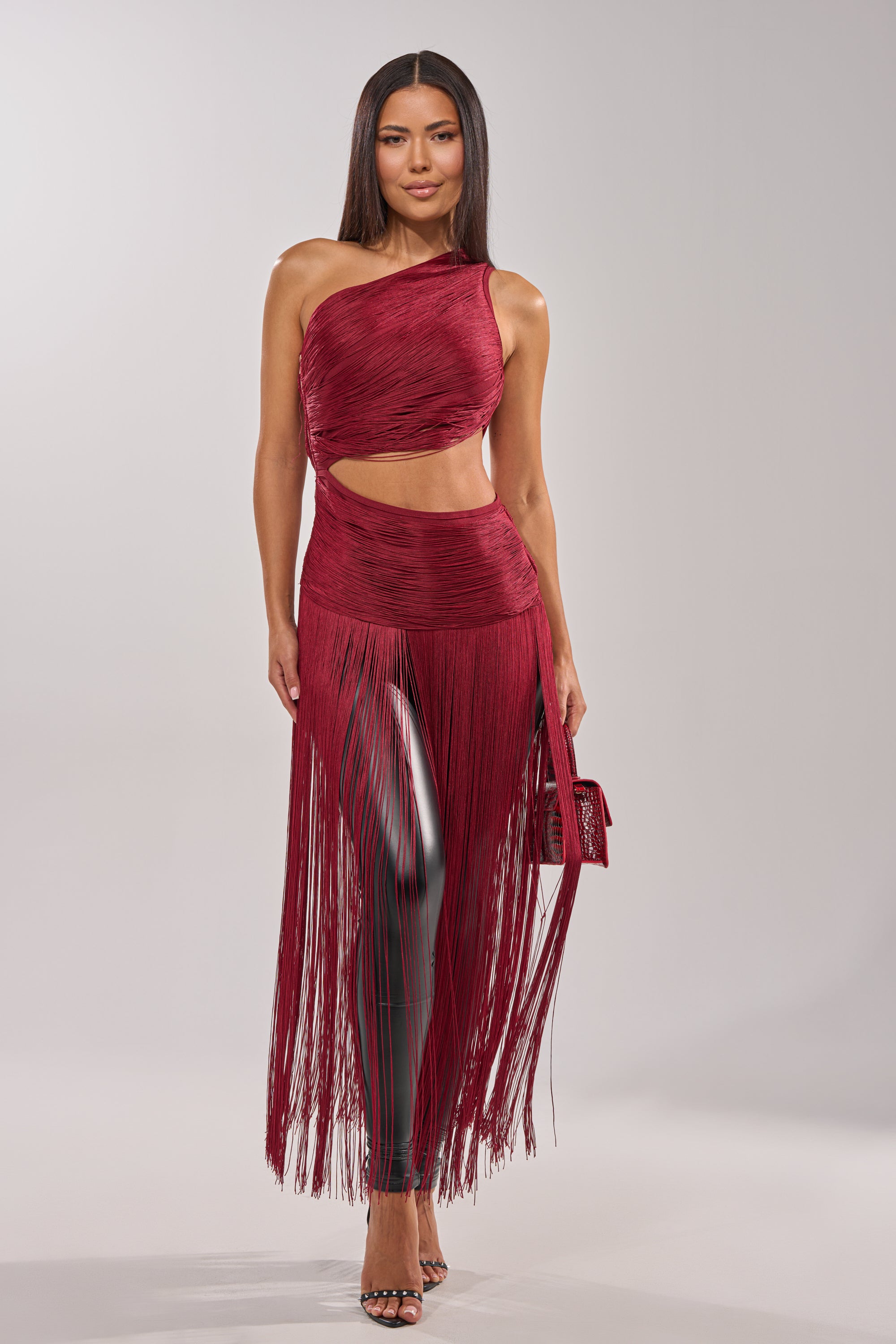A woman stands confidently in the LOVE STRUCK FRINGE TOP, styled as a one-shoulder burgundy crop with fringe and waist cutout. She pairs it with high heels and a matching handbag, her straight hair sleek against a plain light background. 3