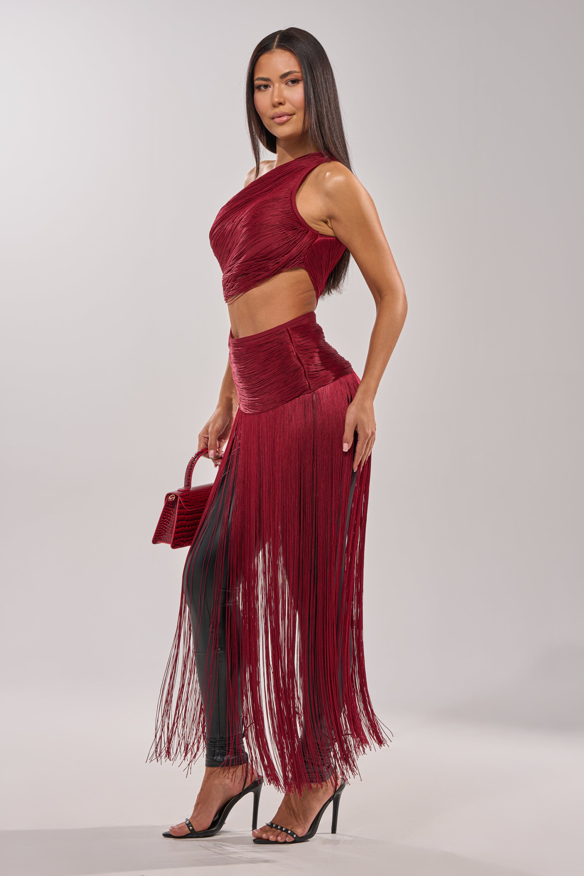 A woman poses against a plain light background wearing the LOVE STRUCK FRINGE TOP in burgundy, styled as a crop top with waist cutouts, paired with a matching red handbag and black high-heeled sandals. 4