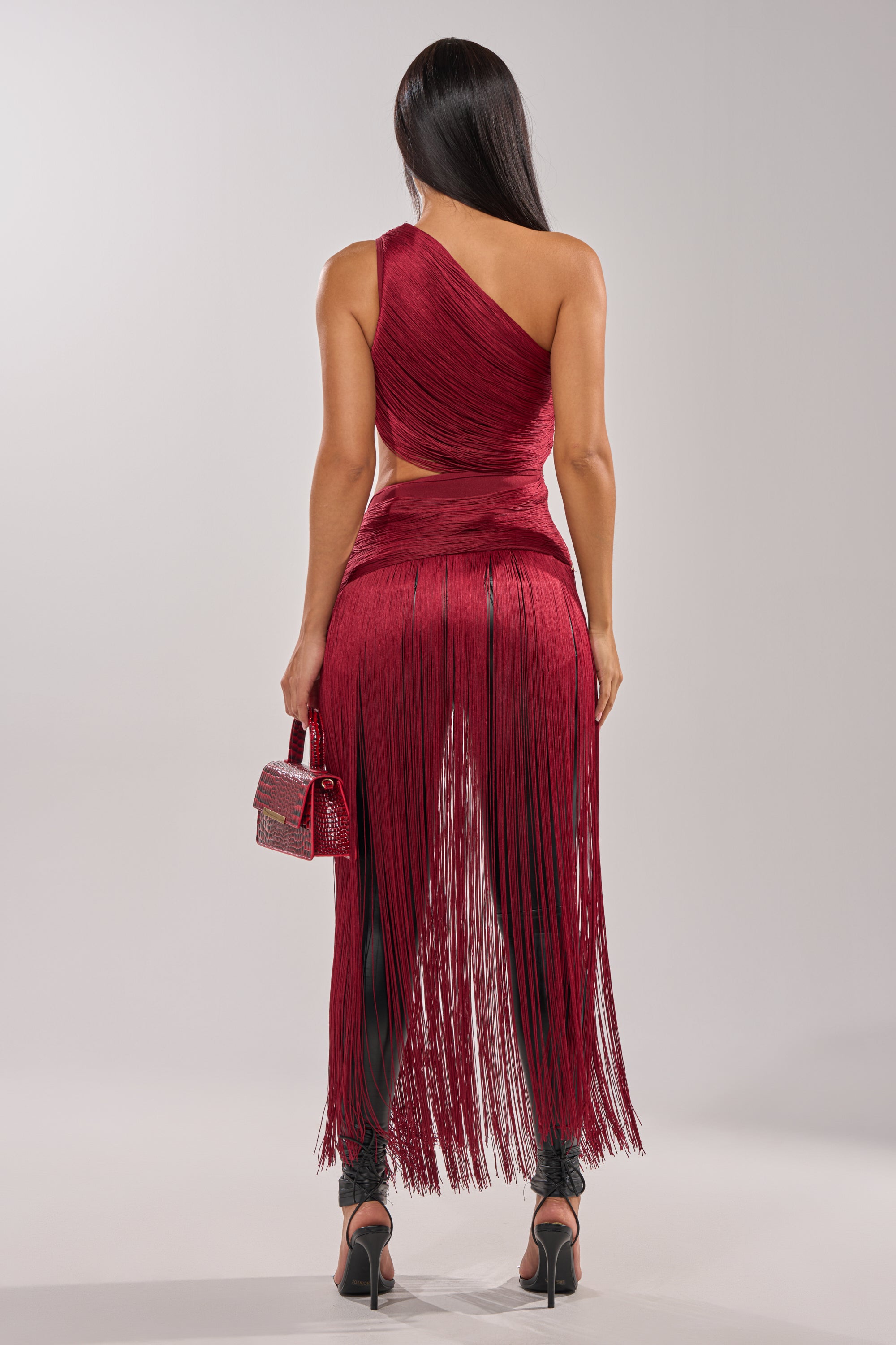 A woman with long, straight dark hair stands against a plain light background, wearing the LOVE STRUCK FRINGE TOP—a one-shoulder burgundy crop top with long fringe—paired with black heels and a small matching red handbag. 6