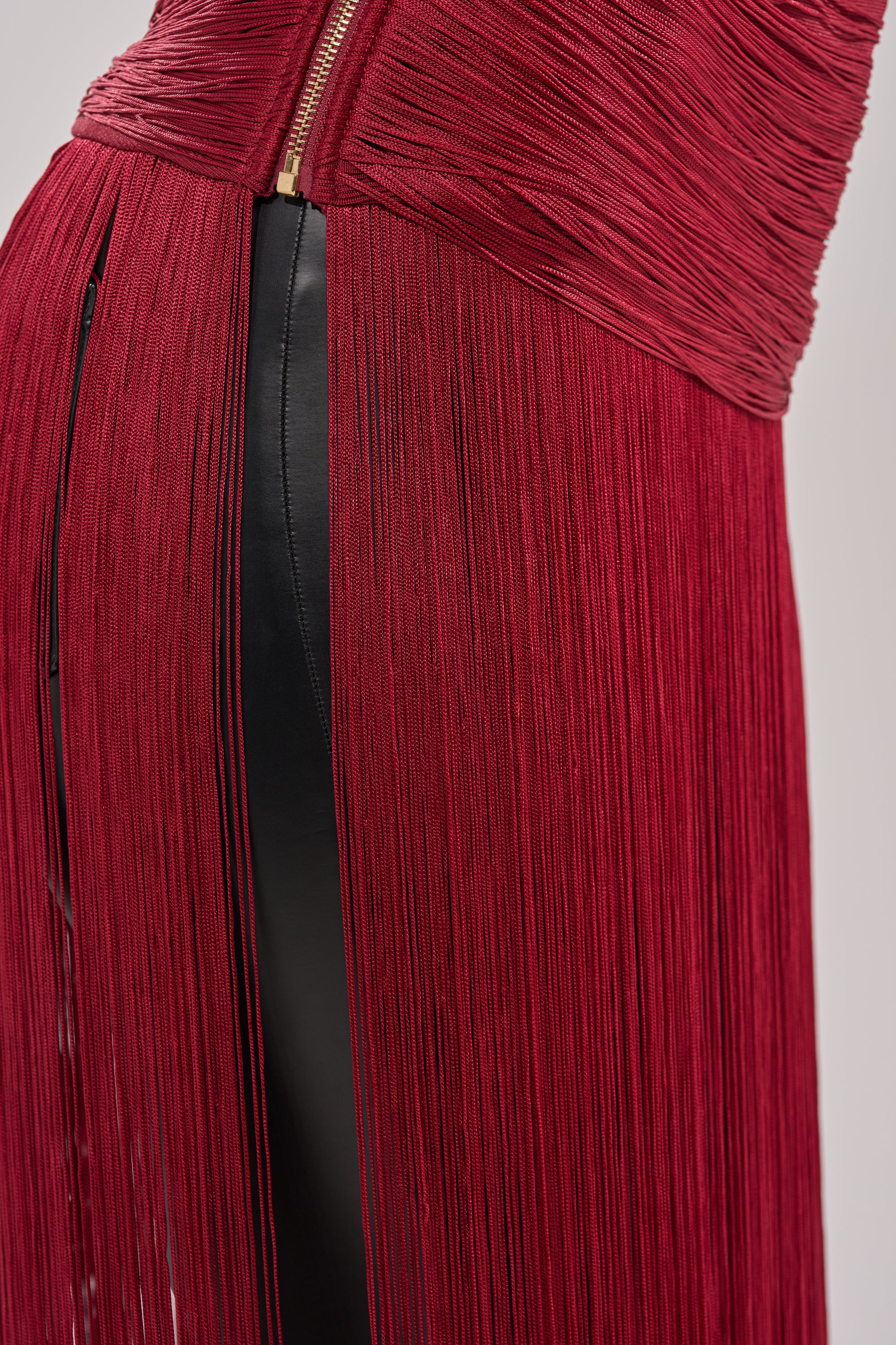 Close-up of the LOVE STRUCK FRINGE TOP: textured, ruched red fabric with long burgundy fringe draping over a sleek black panel, finished with a visible gold zipper at the top. 8