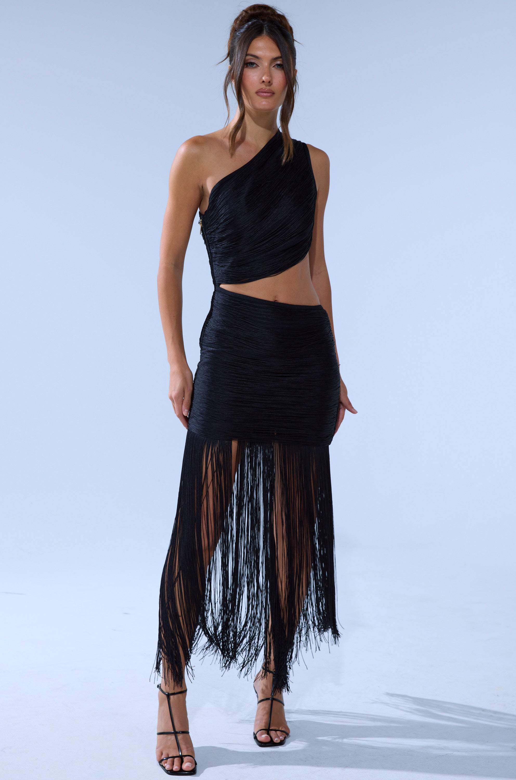 A woman wears the FALLON FRINGE MINI DRESS, a black one-shoulder dress with an asymmetrical crop top and skirt featuring long fringe, paired with strappy black heels against a light background.