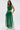 A woman stands against a white background wearing the GINA Single Shoulder Fringe Top in Kelly Green, styled with a long emerald skirt, gold bracelet, and her wavy brown hair cascading down.