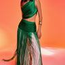 A woman poses against a vibrant orange backdrop, wearing the GINA SINGLE SHOULDER FRINGE TOP IN KELLY GREEN styled over shimmering white pants, and accessorizes with a bold gold cuff bracelet.