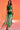 A woman poses before a warm orange backdrop in the GINA SINGLE SHOULDER FRINGE TOP IN KELLY GREEN, a one-shoulder top with fringe, paired with a flowing skirt that reveals a white underskirt. She accessorizes with a gold cuff bracelet.