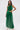 Against a white background, a woman wears the GINA Single Shoulder Fringe Top in Kelly Green with a cutout waist and matching floor-length skirt, posing confidently with one hand on her hip.