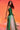 A woman poses confidently against a vibrant orange background, wearing the GINA SINGLE SHOULDER FRINGE TOP IN KELLY GREEN over white pants. Her dark hair is styled in loose waves as she looks elegantly at the camera.