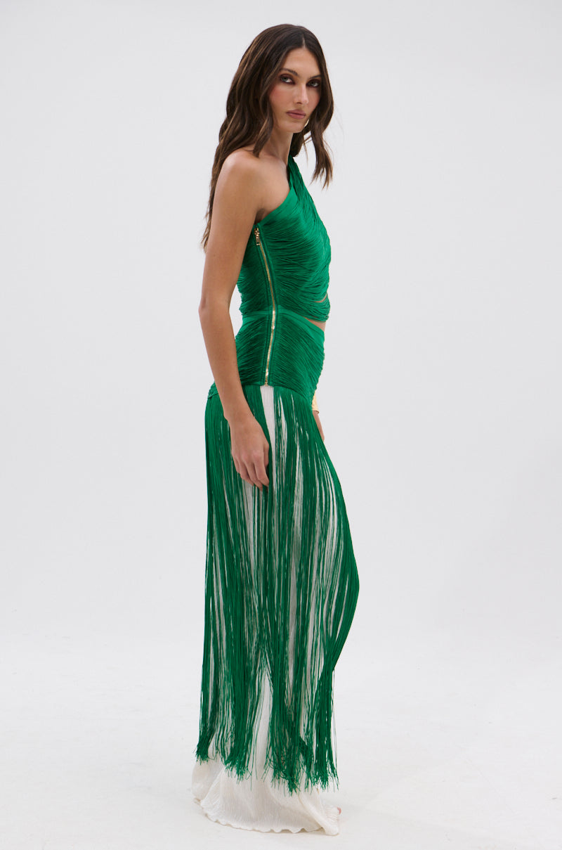 A woman with loose waves stands sideways against a plain white background, wearing the GINA SINGLE SHOULDER FRINGE TOP IN KELLY GREEN over a white underlayer.
