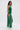 A woman with loose waves stands sideways against a plain white background, wearing the GINA SINGLE SHOULDER FRINGE TOP IN KELLY GREEN over a white underlayer.