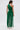 A woman with long, wavy brown hair and gold accessories wears the GINA SINGLE SHOULDER FRINGE TOP IN KELLY GREEN over a white skirt. The fringe drapes from waist to hip, adding texture and flow as she stands facing away.