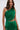 A woman with wavy brown hair wears the GINA Single Shoulder Fringe Top in Kelly Green and a matching skirt, standing against a plain light background and looking confidently at the camera.
