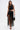 A woman in the GINA SINGLE SHOULDER FRINGE TOP and a matching high-waisted skirt with long fringe details, holding a black handbag and heels, walks confidently against a plain white background.