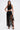 A woman stands confidently in the GINA SINGLE SHOULDER FRINGE TOP, a black sleeveless asymmetrical crop with long fringe, paired with a matching high-waisted skirt. She accessorizes with a black handbag and strappy heels against a plain white background.