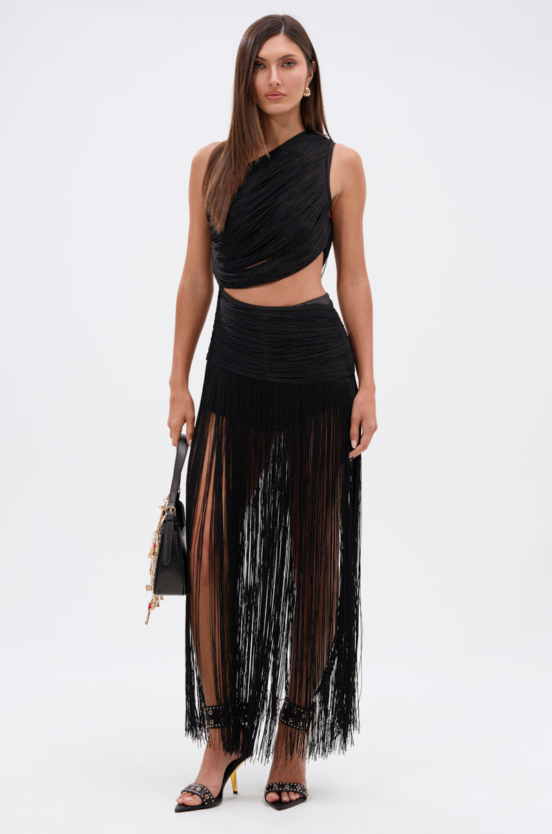 A woman poses against a plain background wearing the GINA SINGLE SHOULDER FRINGE TOP, featuring an asymmetric cut-out and stretch knit bodice. She pairs it with a long black fringe skirt, black handbag, and high-heeled sandals.