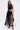 A woman with long brown hair wears the GINA SINGLE SHOULDER FRINGE TOP and a high-waisted, long black fringe skirt. She holds a black handbag and wears embellished high-heeled sandals against a plain white background.
