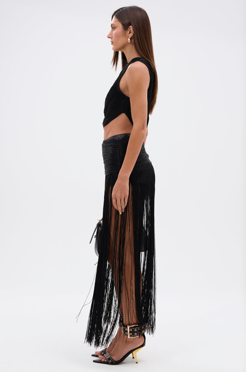 A woman in profile wears the GINA SINGLE SHOULDER FRINGE TOP with a high-waisted black skirt featuring long fringe, black heels with yellow accents, and carries a small handbag—an elegant floor-grazing showstopper look.