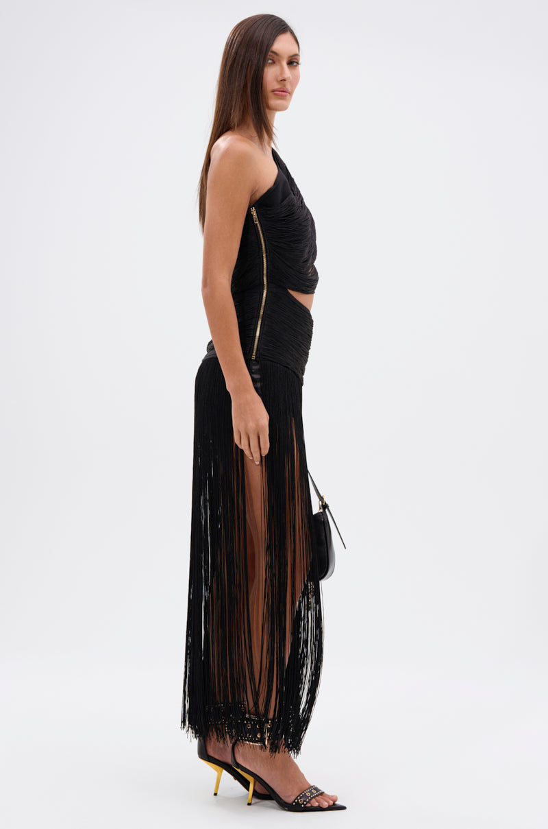 A woman stands in profile against a white background wearing the GINA SINGLE SHOULDER FRINGE TOP and a black fringed skirt. She accessorizes with a small black purse and high-heeled sandals, embodying showstopping style.