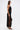 A woman stands in profile against a white background wearing the GINA SINGLE SHOULDER FRINGE TOP and a black fringed skirt. She accessorizes with a small black purse and high-heeled sandals, embodying showstopping style.