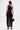 A woman faces away, wearing the GINA SINGLE SHOULDER FRINGE TOP—a black, one-shoulder top with long fringe details. She pairs it with a black handbag and high heels, standing against a plain white background.
